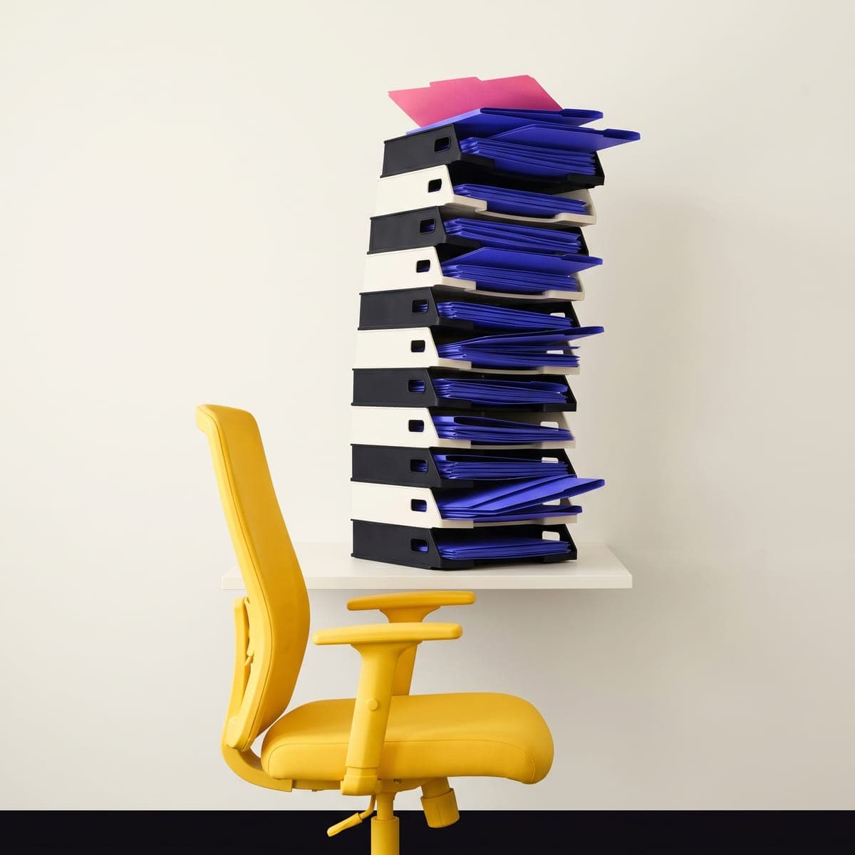 Tour de bannettes courrier débordant de dossiers indigo, posée sur un bureau au-dessus d’une chaise jaune vide.